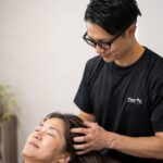 ベッドに仰向けになった女性の頭に手をあて、ヘッドケアを行うトレーナー